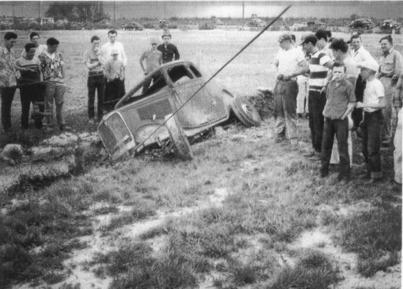 A racing car is being pulled out of a ditch as a crowd looks on.